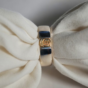 May include: A silver ring with a gold spiral design in the center. The ring is rectangular with a polished finish. The ring is displayed on a white fabric background.