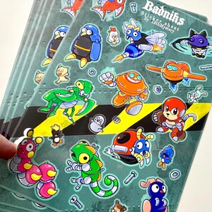 May include: A stack of sticker sheets featuring various cartoon characters in vibrant colors. The stickers include robots, fish, penguins, and other creatures. The text "Badniks" and "Sticker sheet by 12thMystery" are visible.