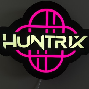 May include: A black sign with the word "HUNTRIX" in a bright, glowing, off-white font. The sign has a pink, geometric design and a black border. The sign is illuminated and appears to be made of plastic.