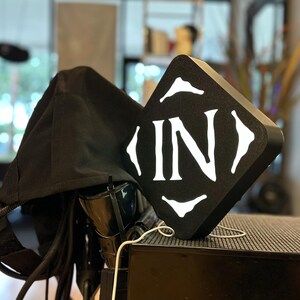 May include: A black square-shaped object with the word "IN" in white, stylized text. The object is placed on a dark surface next to a black fabric hood. A white cable is connected to the object.