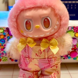 May include: A plush doll in pink and yellow, wearing a hooded head covering and overalls. The doll features large, round eyes, a small yellow beak, and a toothy grin. The overalls are patterned with the word "love". Two yellow bows are on the chest.