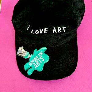 Könnte beinhalten: Schwarze Baseballkappe mit weißer Stickerei, die "I LOVE ART" sagt. Ein grüner Patch mit weißer Stickerei, der "SUPPORT THE ARTS" sagt, ist an der Vorderseite der Kappe befestigt.
