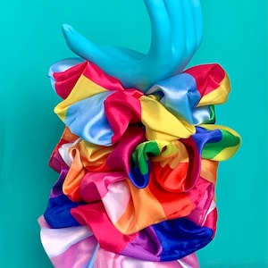 Pode incluir: Uma colorida e vibrante scrunchie de cetim com as cores do arco-íris é exibida em um suporte em forma de mão azul claro. A scrunchie apresenta uma variedade de cores, incluindo rosa, amarelo, laranja e roxo, criando um acessório ousado.