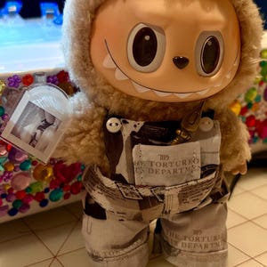 May include: A soft toy doll with a smiling face, wearing a furry hood and printed overalls. The overalls have a small pocket and text. The doll is holding a small, clear framed picture. The doll's skin is a light tan colour.