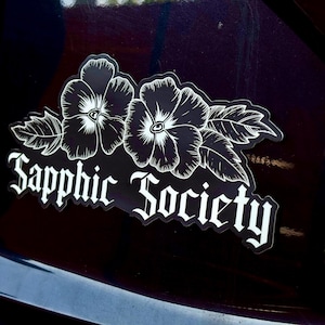 Puede incluir: Una pegatina en blanco y negro con dos flores estilizadas y la frase "Sapphic Society" en una fuente gótica. La pegatina está diseñada para un coche u otra superficie similar.