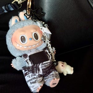 May include: A novelty keychain with a cartoon character in a gray hooded outfit and overalls printed with text. The keychain includes a small white figure and a chain with charms, including an 8-ball. The text on the overalls reads "I did something bad".