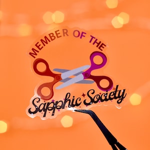Könnte beinhalten: Ein transparenter Aufkleber mit einer pinkfarbenen und roten Schere, dem Text "Member of the Sapphic Society" mit Glitzerakzenten und einem schwarzen Hintergrund.