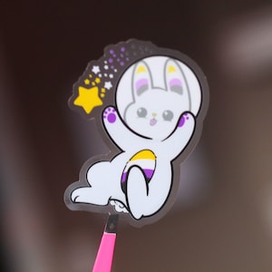 Pode incluir: Um coelho branco vestindo um traje espacial com uma estrela amarela e listras roxas, amarelas e brancas no peito. O coelho está flutuando no espaço com um rastro de estrelas atrás dele.