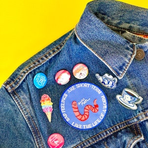 May include: A blue denim jacket with various embroidered patches and pins. The patches include a worm, a teacup, a ice cream cone, and a few others. The text on one of the patches reads "Sad Feelings Are Short-Term... Like The Life Of A Worm".