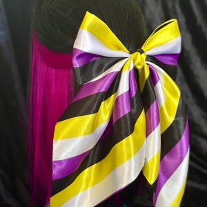 Peut inclure: Un grand nœud en satin aux couleurs de la fierté non binaire : rayures jaunes, blanches, violettes et noires. Le nœud est noué à l'arrière d'une chevelure avec des cheveux noirs et une mèche rose vif. Accessoire pour cheveux.