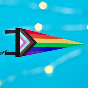 May include: A rainbow pride flag pennant with a brown, pink, light blue, and white triangle in the upper left corner. The pennant is made of clear vinyl and has a white string attached to the top.