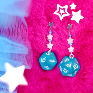 Può includere: Un paio di orecchini pendenti con ciondoli a forma di dado blu e perline a forma di stella bianca. Gli orecchini sono su uno sfondo rosa peloso.