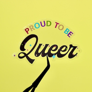 Puede incluir: Una pegatina con el texto "Proud to be Queer" en una fuente arcoíris. La pegatina está sobre un fondo amarillo y tiene un efecto de purpurina arcoíris.