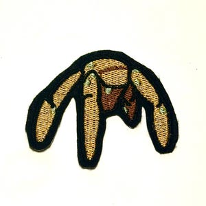 Puede incluir: Parche bordado con un diseño abstracto. Tiene un borde negro, con un interior marrón y beige. El diseño parece una mano o garra estilizada. El parche está diseñado para ser cosido o planchado en ropa o accesorios.
