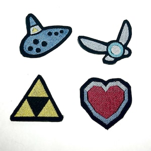 May include: A set of four embroidered patches featuring a blue ocarina with black dots, a small winged fairy in light blue and white, a yellow triangle with a black centre, and a red heart shape outlined in light blue and dark blue borders.