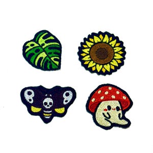 May include: Four embroidered patches: a green leaf with black outlines, a yellow sunflower with a brown centre, a black moth with a skull design, and a mushroom with a red cap and smiling face. All patches have black borders.