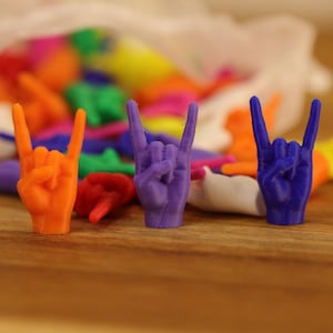 May include: A collection of small, three-dimensional plastic hand figurines shaped into the rock and roll sign. The set includes various bright colors like orange, purple, and blue, with several pieces spilling out from a sheer white drawstring bag.