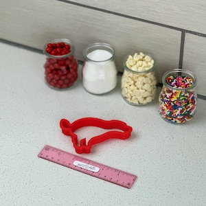 May include: A red pig-shaped cookie cutter with a pink ruler beside it. Four small glass jars filled with red sweets, white sugar, white chocolate chips, and colourful sprinkles are in the background.