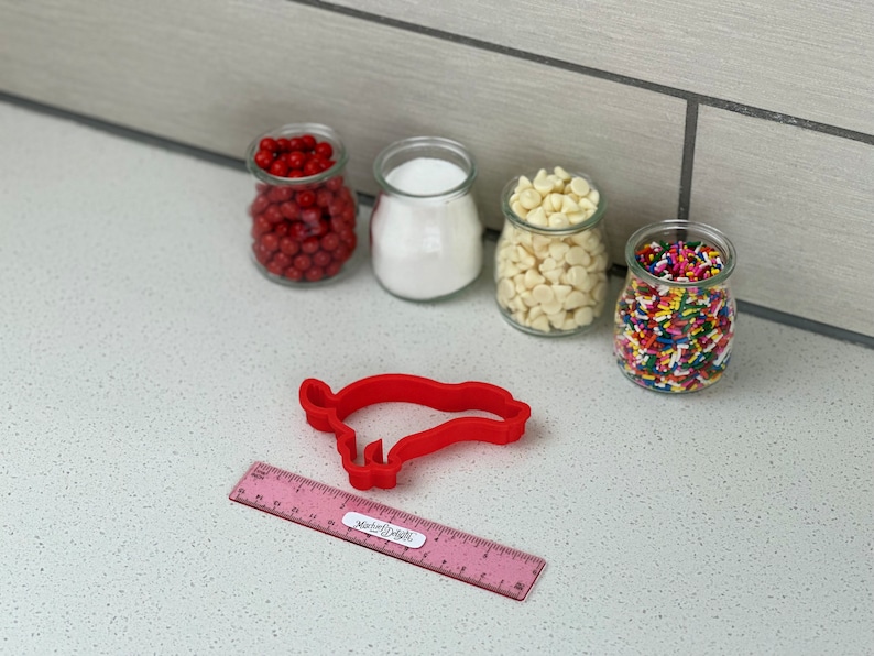 May include: A red pig-shaped cookie cutter with a pink ruler beside it. Four small glass jars filled with red sweets, white sugar, white chocolate chips, and colourful sprinkles are in the background.