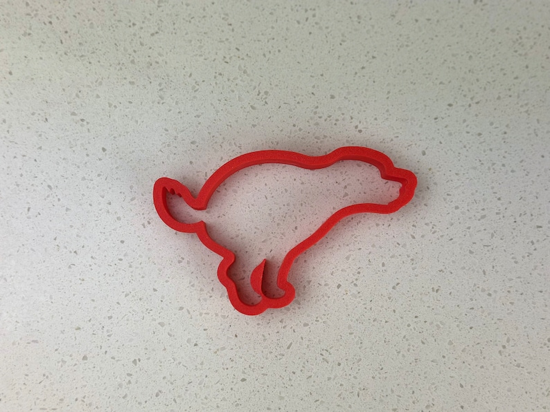 May include: A red plastic cookie cutter shaped like a running dog. The cutter's outline defines the dog's form, including its legs, tail, and body. The design is simple and clean, suitable for baking.