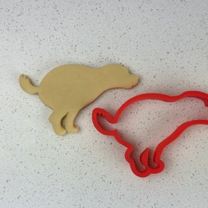 May include: A tan dog-shaped biscuit and a red dog-shaped biscuit cutter are shown on a white speckled surface. The cutter is a single piece of red plastic, and the biscuit is a light tan colour.