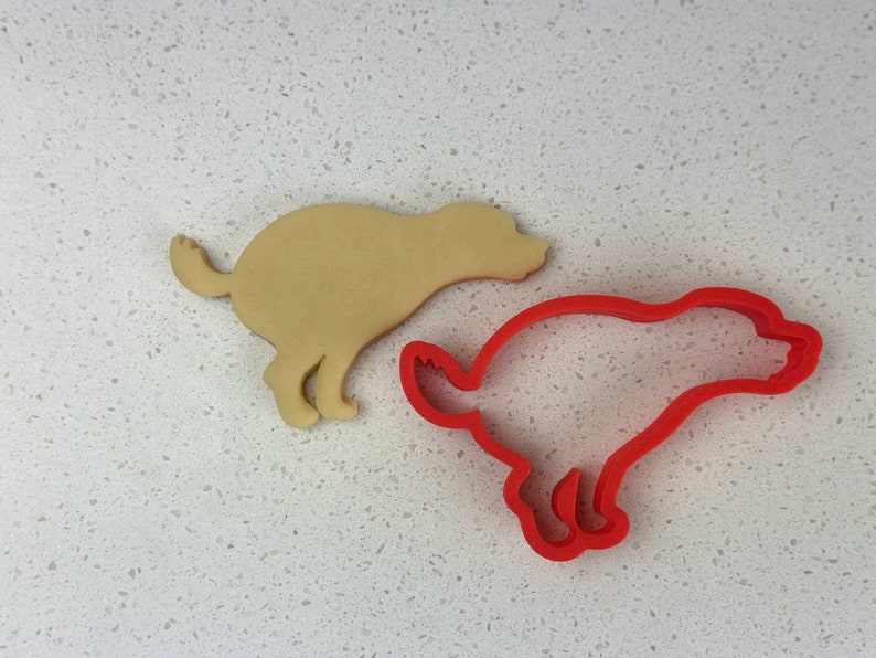 May include: A tan dog-shaped biscuit and a red dog-shaped biscuit cutter are shown on a white speckled surface. The cutter is a single piece of red plastic, and the biscuit is a light tan colour.