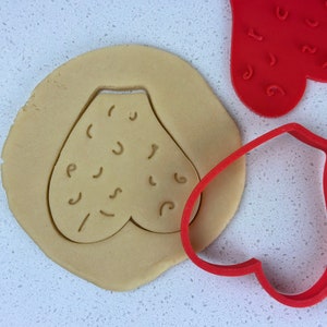 May include: Red heart-shaped cookie cutter with a pattern of small lines and squiggles. A second red heart-shaped cookie cutter is shown on top of a piece of cookie dough, with the cutter pressed into the dough.