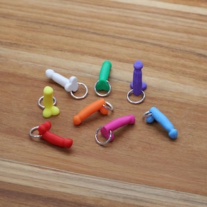 May include: A collection of colorful penis-shaped keychains in various colors, including red, orange, pink, green, blue, yellow, and purple. Each keychain has a silver ring attached. The keychains are arranged on a wooden surface.