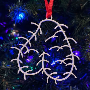 May include: White, heart-shaped Christmas ornament with a delicate, intricate design resembling branches. The ornament is hanging from a red ribbon on a Christmas tree.