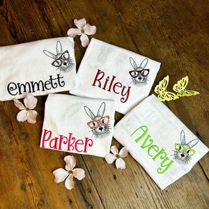 May include: Four white t-shirts with embroidered images of bunnies wearing glasses and personalized names: Emmett, Riley, Parker, and Avery.