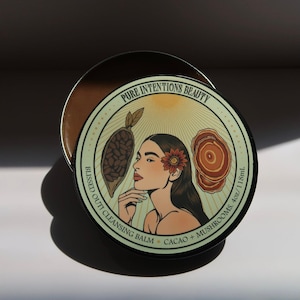 May include: A round, open container of cleansing balm with a decorative lid. The lid features the text "PURE INTENTIONS BEAUTY" and "BLISSED OUT! CLEANSING BALM CACAO + MUSHROOMS. 4oz/118mL". The lid's design includes a woman's portrait, cacao, and mushrooms.