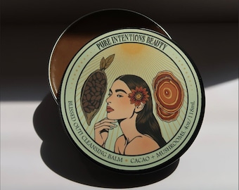 May include: A round, open container of cleansing balm with a decorative lid. The lid features the text "PURE INTENTIONS BEAUTY" and "BLISSED OUT! CLEANSING BALM CACAO + MUSHROOMS. 4oz/118mL". The lid's design includes a woman's portrait, cacao, and mushrooms.