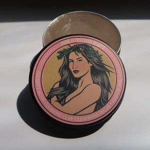 May include: A round, black tin of acne clearing clay cleansing balm. The lid features a woman with long dark hair and a floral crown. Text on the lid reads "Pure Intentions Beauty" and "Lucidity Acne Clearing Clay Cleansing Balm". The balm is 4 oz / 118.3mL.