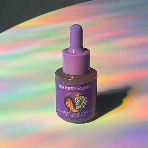 May include: A small, clear glass bottle with a purple dropper and cap. The label reads "Pure Intentions Beauty" and "Mangosteen Dreams Oil Serum". The bottle is set against a background with rainbow light reflections.