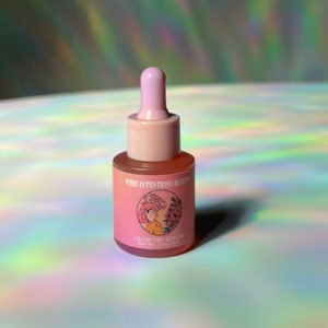 Puede incluir: Una peque&ntilde;a botella de vidrio rosa de Glow Oil Serum con cuentagotas. La botella tiene una tapa rosa y una etiqueta con el texto "Pure Intentions Beauty" y una ilustraci&oacute;n floral. El fondo es una superficie brillante e iridiscente.