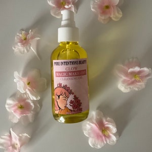 May include: A clear glass bottle filled with yellow liquid, labeled "Pure Intentions Beauty Glow Magic Make-Off Cleansing Oil." The label features a pink background with an illustration of a woman and pink flowers. The bottle is surrounded by pink and white artificial flowers.