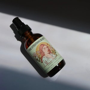 May include: A brown glass spray bottle with a black nozzle. The label reads "Pure Intentions Beauty" and features an illustration of a woman. The bottle contains "Ethereal Mist" with honeysuckle, peach, and mask ingredients. Net wt. 2 oz/60ml.