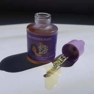 May include: A clear glass bottle of oil serum with a purple label that reads "Pure Intentions Beauty" and "Soften Dreams Oil Serum". The bottle is open, with a light purple dropper next to it. The serum is a light amber color.