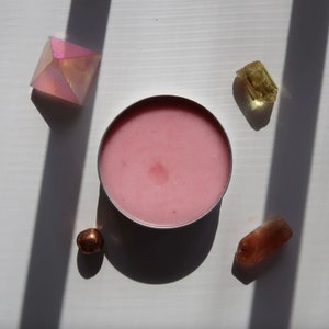 May include: A pink cosmetic product in a silver container is surrounded by crystals and a small metallic sphere. The crystals are various colors, including pink, yellow, and orange. The objects are arranged on a white surface with shadows.