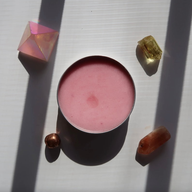 May include: A pink cosmetic product in a silver container is surrounded by crystals and a small metallic sphere. The crystals are various colors, including pink, yellow, and orange. The objects are arranged on a white surface with shadows.