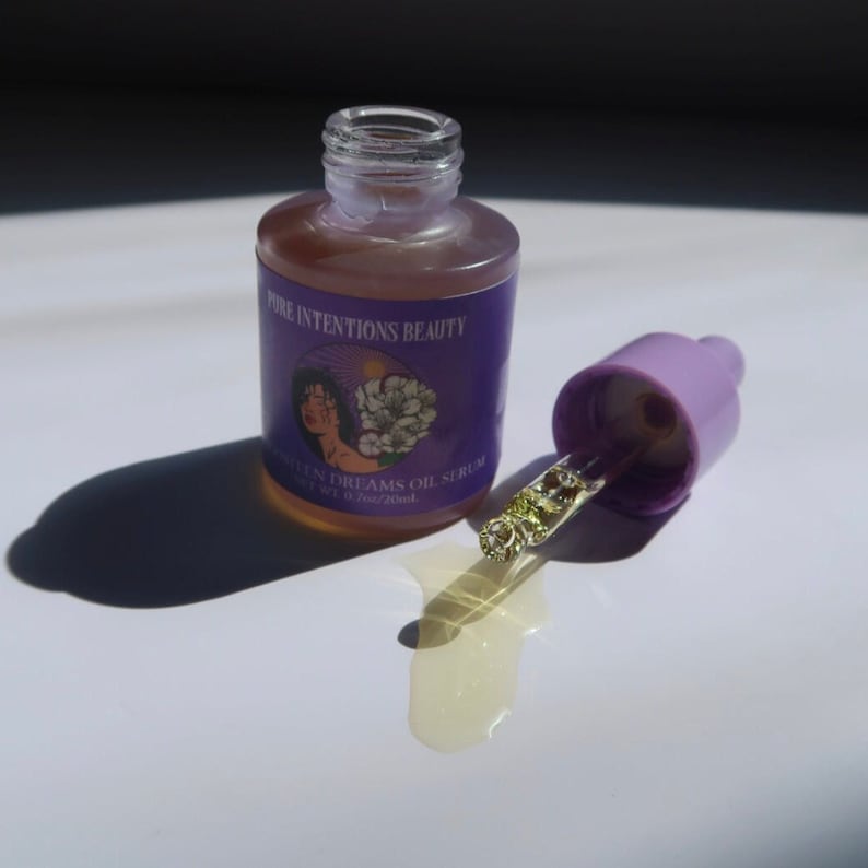 May include: A clear glass bottle of oil serum with a purple label that reads "Pure Intentions Beauty" and "Soften Dreams Oil Serum". The bottle is open, with a light purple dropper next to it. The serum is a light amber color.