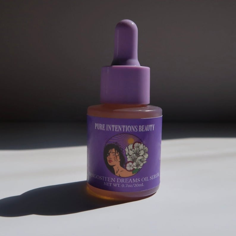 May include: A small, clear glass bottle of Mangosteen Dreams Oil Serum with a purple dropper and cap. The label features the text "Pure Intentions Beauty" and a graphic of a woman and flowers. The bottle contains 0.7 oz/20ml of serum.