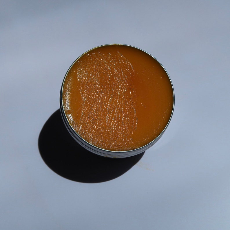 May include: A round, open metal tin filled with a smooth, amber-colored balm. The balm's surface has visible texture, and the tin casts a dark shadow on the white surface below.