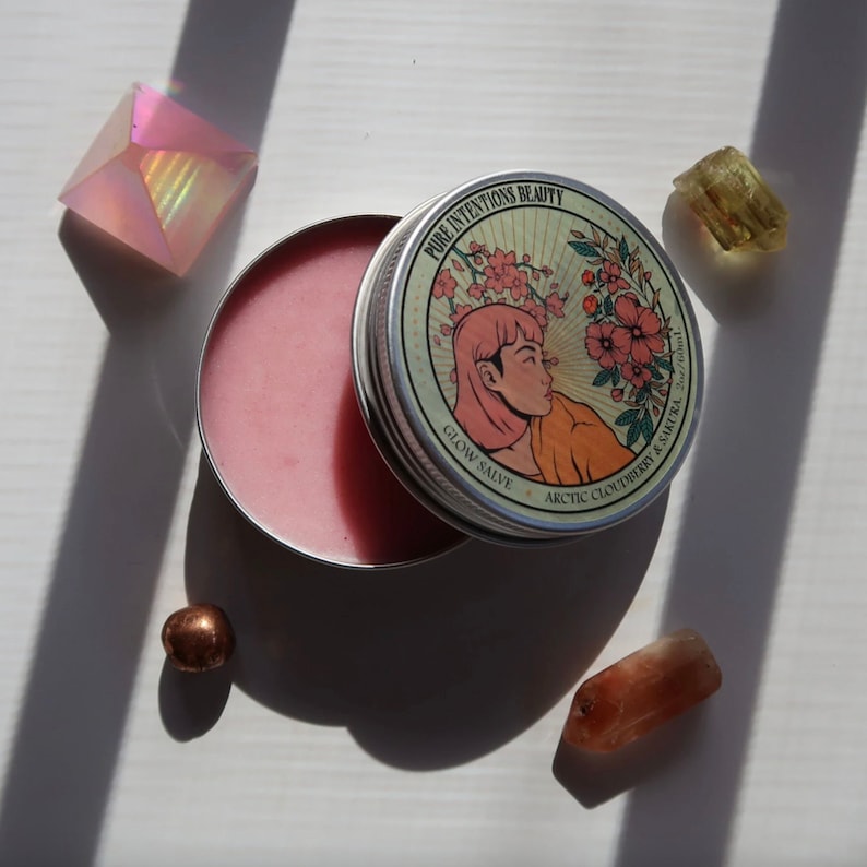 May include: An open, round metal tin of pink Glow Salve with the text "Pure Intentions Beauty" and "Arctic Cloudberry & Sakura." The tin's lid features a woman's profile with flowers. The image includes crystals and a small copper-colored sphere.