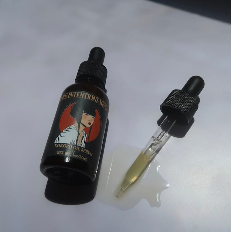May include: A brown glass bottle of Kokoro Oil Serum with a black dropper and a label featuring a woman's portrait. The dropper is next to the bottle, with a small amount of oil spilled out. The bottle reads "Pure Intentions Beauty".