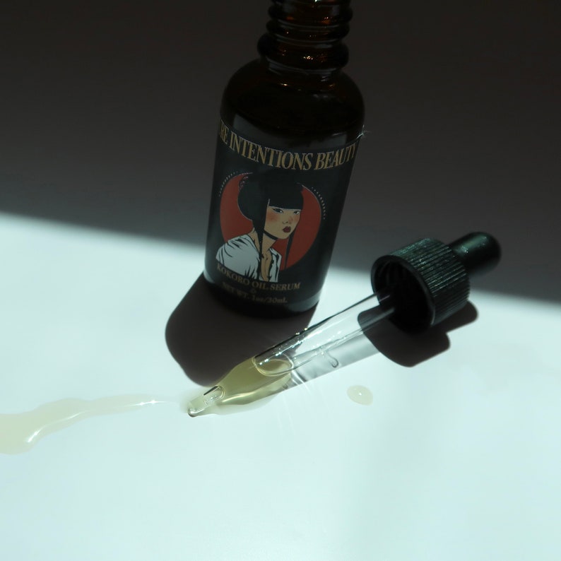 May include: A dark brown glass bottle of Kokoro Oil Serum with a black dropper. The label features a woman's portrait in a red circle. A small amount of serum is spilled on the white surface.