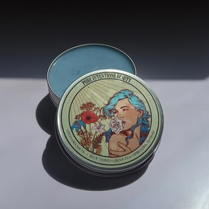 May include: An open, round metal tin with a light blue balm inside. The lid features an illustration of a woman with blue hair and a flower in her mouth, surrounded by text that reads "PURE INTENTIONS BEAUTY" and "ATHANASIA SALVE BLUE TANSY+GREEN TEA+SPIRULINA. 2oz/60ml."
