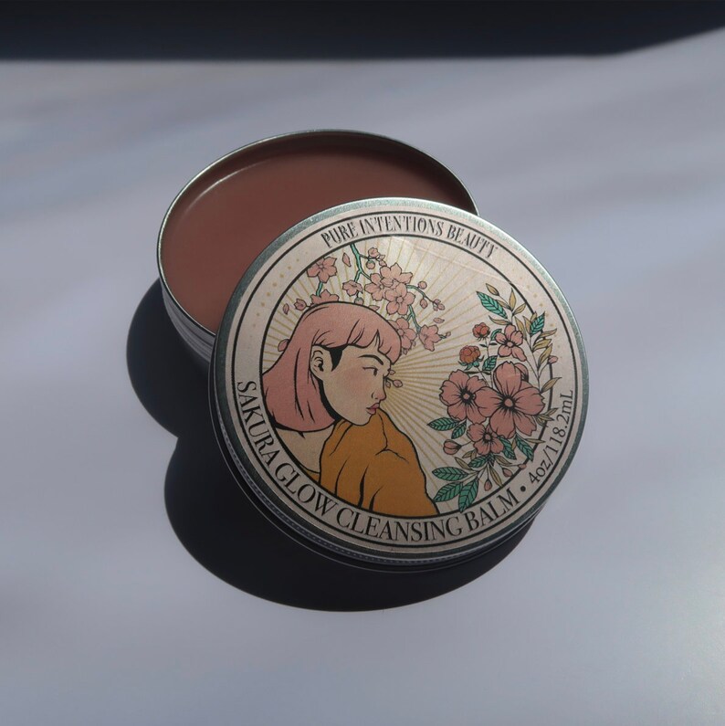 May include: Open metal tin of Sakura Glow Cleansing Balm. The lid features a woman with pink hair and floral accents. The balm is a light brown color. The tin is labeled "Pure Intentions Beauty" and contains 4oz/118.2mL of product.