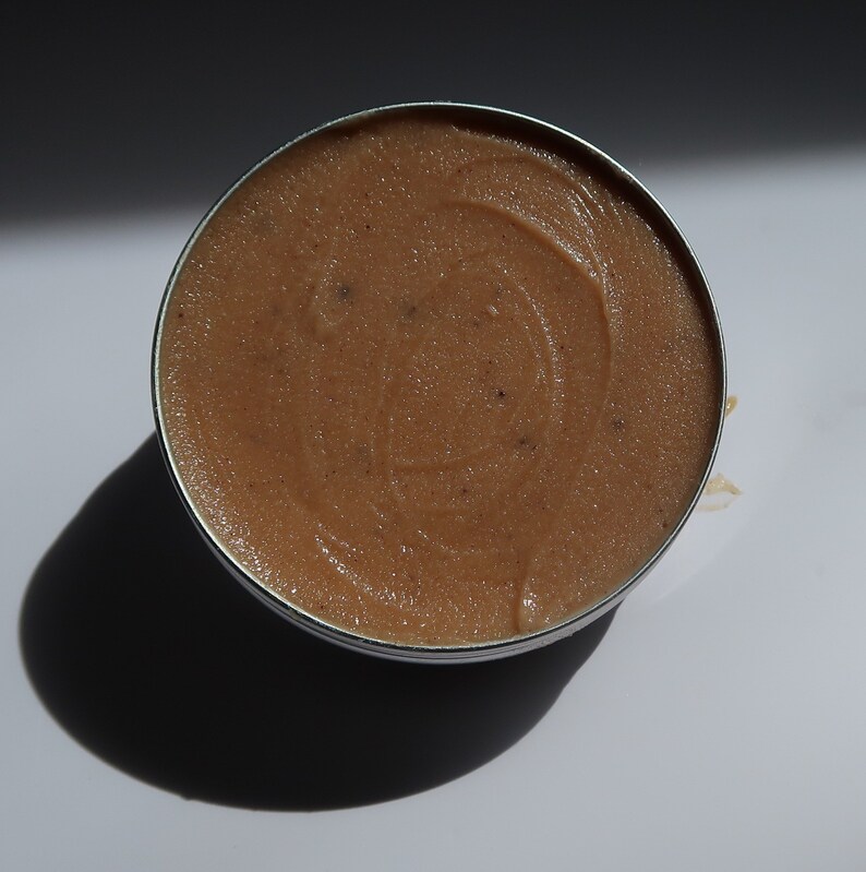 May include: A circular metal tin containing a light brown, textured product. The substance appears smooth and creamy, with tiny dark particles. The tin is on a white surface, creating a dark shadow.