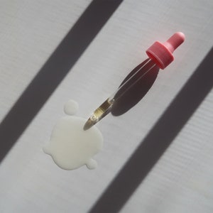 Puede incluir: Primer plano de un cuentagotas de vidrio transparente con una tapa rosa, lleno de l&iacute;quido dorado. Un peque&ntilde;o charco del mismo l&iacute;quido est&aacute; sobre una superficie blanca, con sombras.